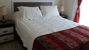 A hotel room with a white brick wall, a wooden headboard, two bedside tables with lamps, a bed with white bedding, and a red and gray patterned throw blanket. - New apartment, 1A center of Villarrica (Villarrica, Araucania)