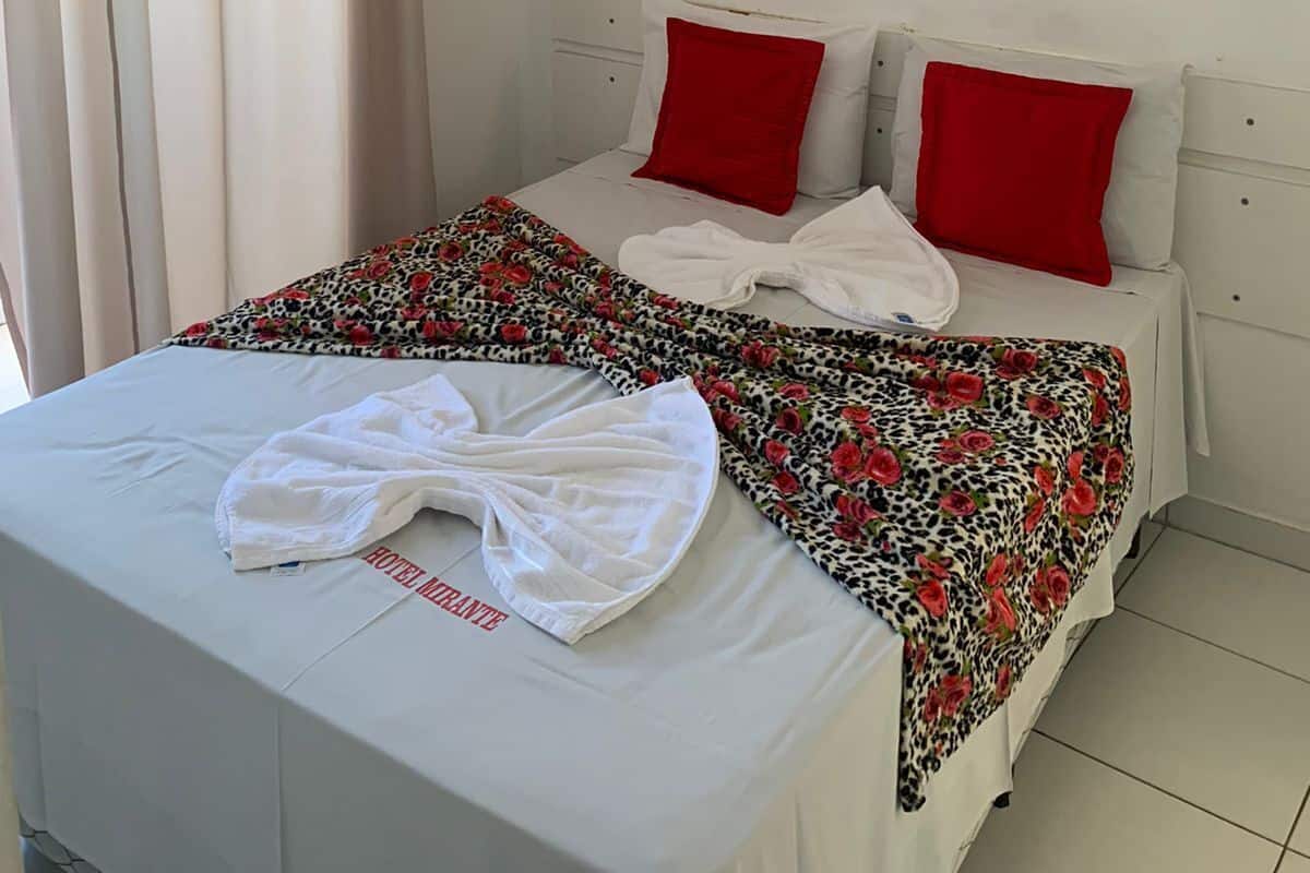 A hotel room with a bed, red pillows, a floral painting, and an air conditioning unit.