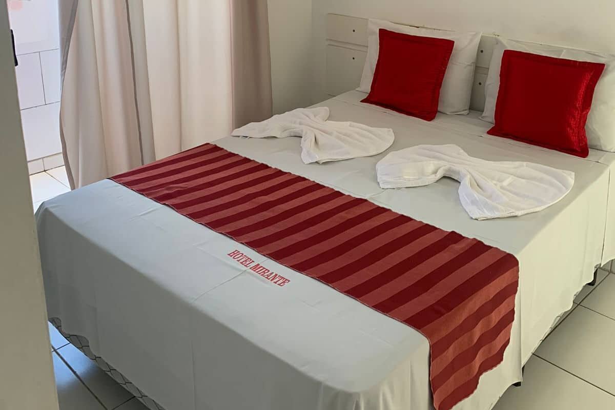 A hotel room with a bed, red pillows, a white blanket, a red-striped rug, an air conditioning unit, and a painting on the wall.