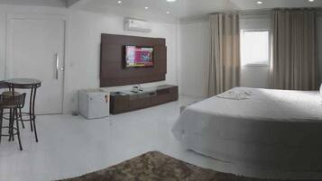 A bedroom with a bed, a TV mounted on the wall, a desk with a chair, and a window with curtains.