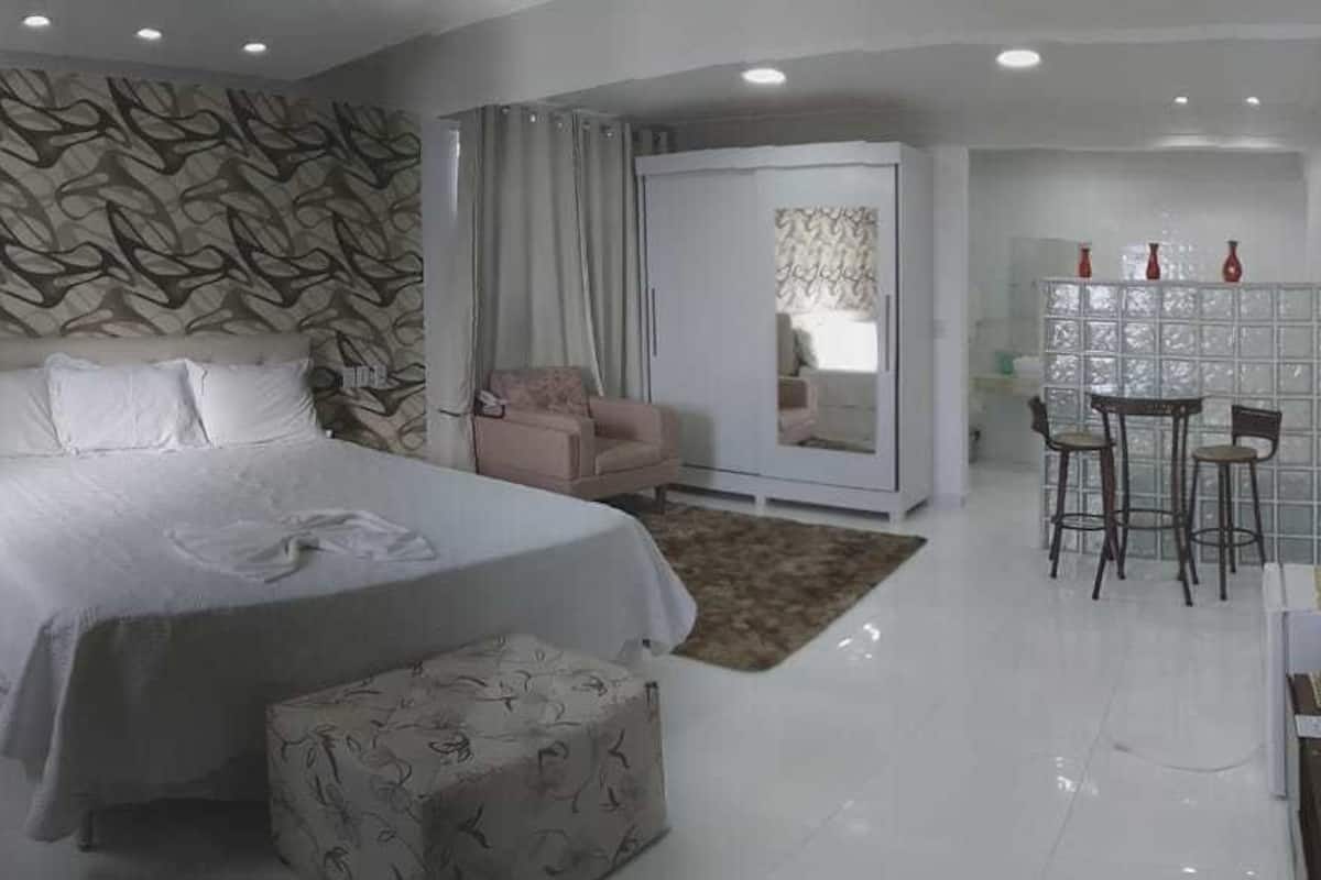 A modern hotel room with a large bed, a seating area with a table and chairs, a wardrobe, and a bathroom visible through a glass door.