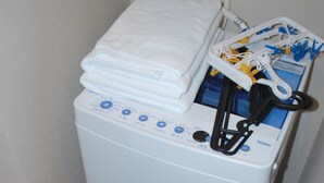 A compact, white, front-loading washing machine with a control panel, a white towel on top, and a black basket at the base. - Ten Point Hotel (Naha)