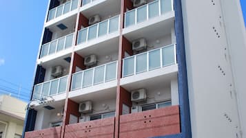 Um edifício de vários andares com design moderno, com varandas em cada pavimento, unidades de ar condicionado e entrada no nível da rua com um carro estacionado na frente.