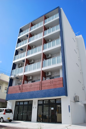 Un edificio de varios pisos con diseño moderno, que cuenta con balcones en cada nivel, unidades de aire acondicionado y una entrada a pie de calle con un coche estacionado afuera.