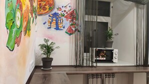 Una habitación con una mesa de madera, una planta en maceta, una pared con arte colorido, una ventana con cortinas y un televisor sobre un mueble.