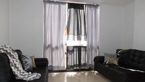 A modern living room with two black sofas, one with patterned throw pillows, and a large window with sheer curtains. - Edificio Angelo (Cartago)