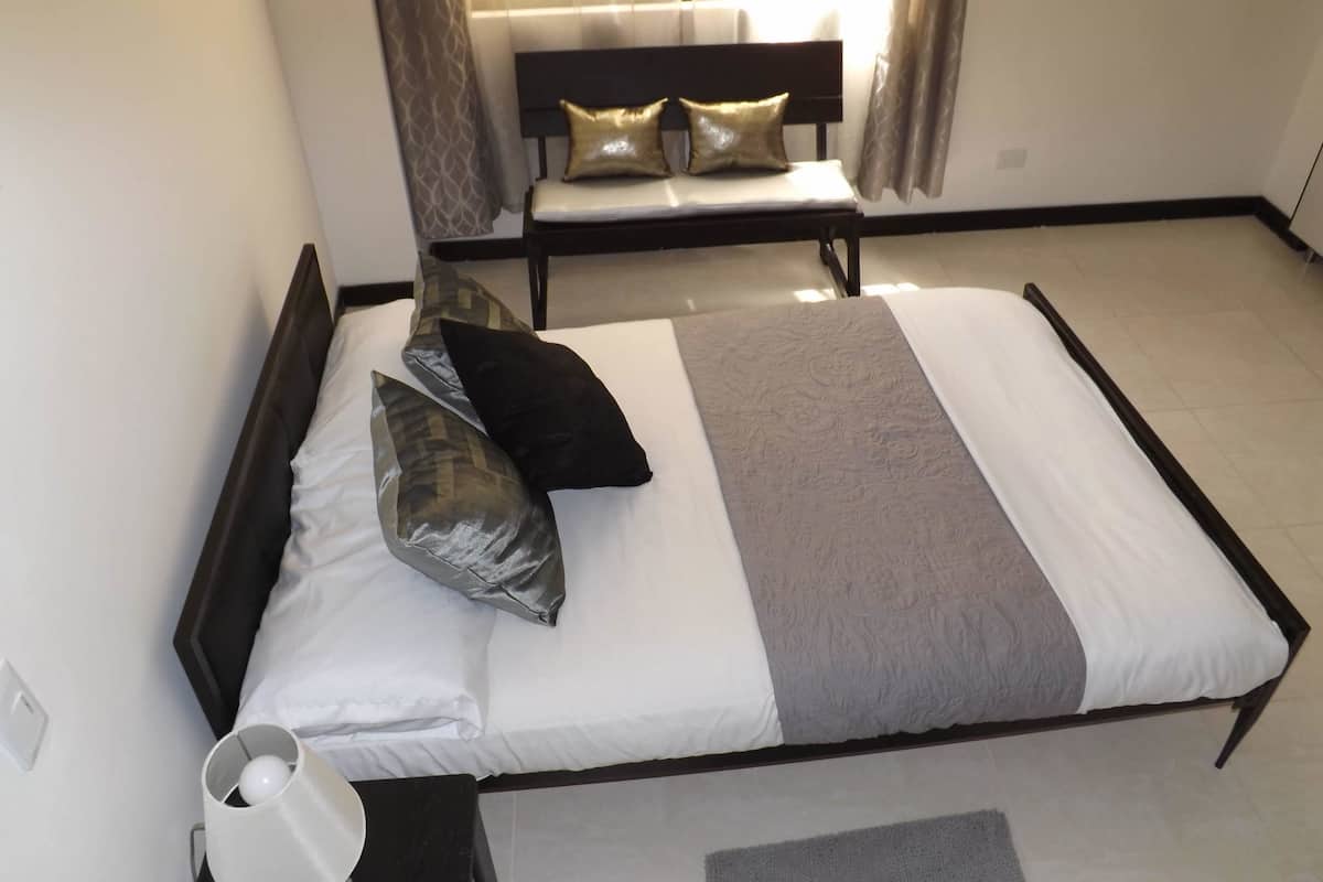 A neatly made bed with a headboard, a bedside table with a lamp, and a bench with cushions in a room with a tiled floor.