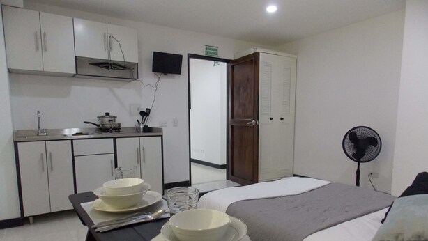 A hotel room with a bed, bedside table with plates and cutlery, a small kitchenette, a fan, and a door with a sign. - Edificio Angelo (Cartago)