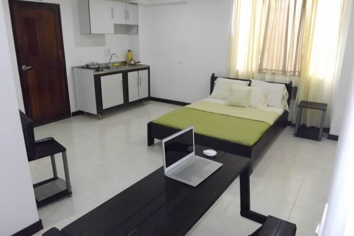 A hotel room with a bed, a desk with a laptop, a small table, a chair, a kitchenette, and a window with curtains.