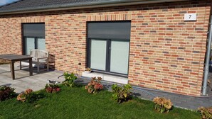Un bâtiment en briques avec deux volets roulants fermés, une enseigne portant le chiffre « 7 », un banc et un jardin fleuri.