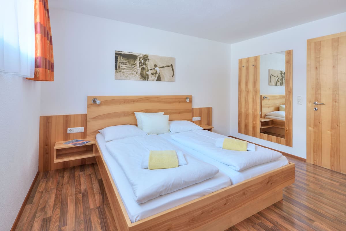 Uma cama de casal com cabeceira e peseira de madeira, uma mesinha de cabeceira, um espelho e um guarda-roupa de madeira.