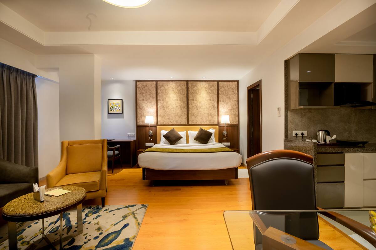 A hotel room with a bed, two chairs, a small table, a kitchen area, and a dining table.