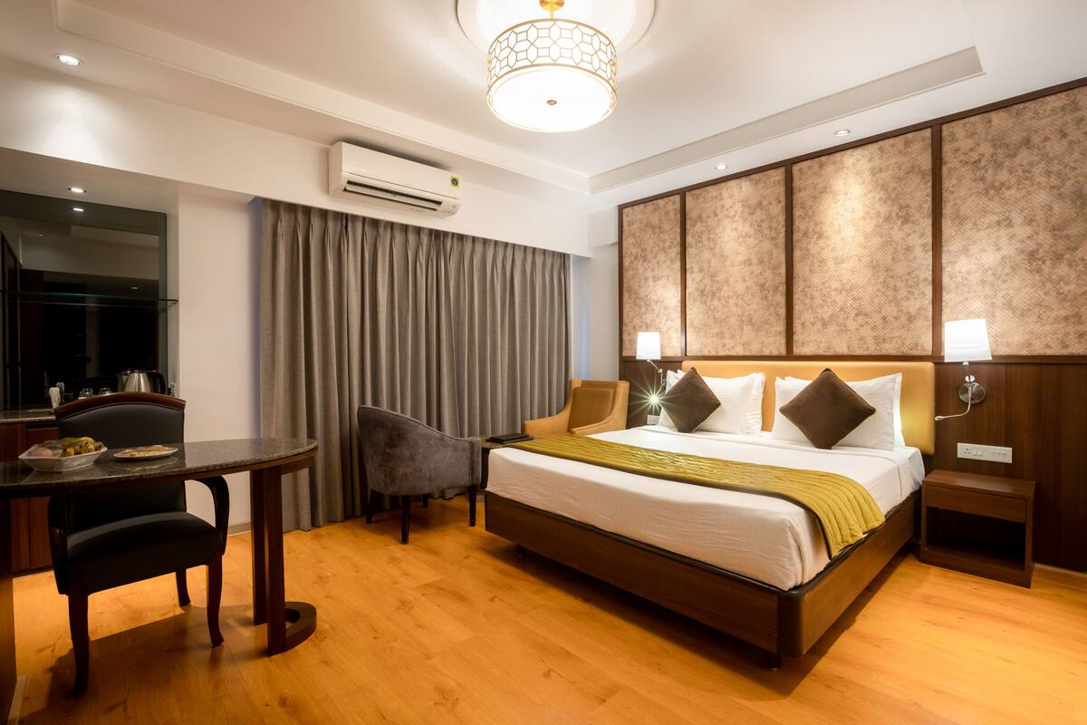 A hotel room with a bed, bedside table, chair, and a desk with a lamp.