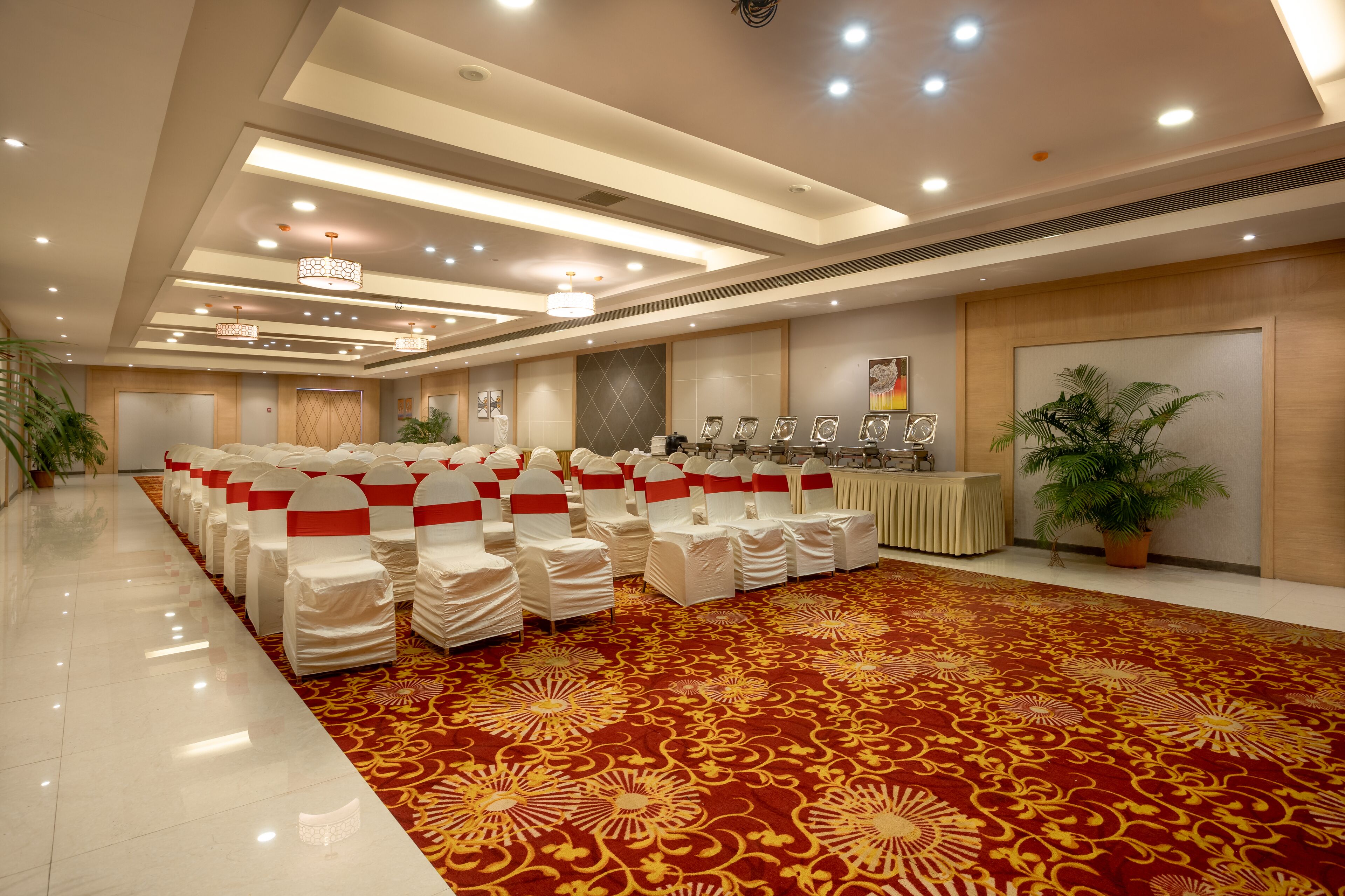 A banquet hall with rows of white chairs with red sashes, a red patterned carpet, and a ceiling with recessed lighting.