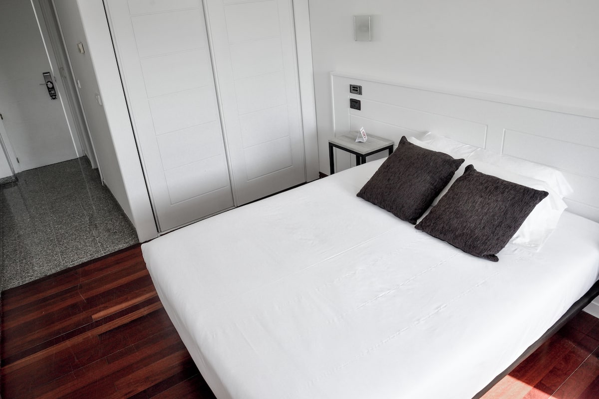 Quarto de hotel com uma cama grande, uma mesa de cabeceira e uma porta com puxador.