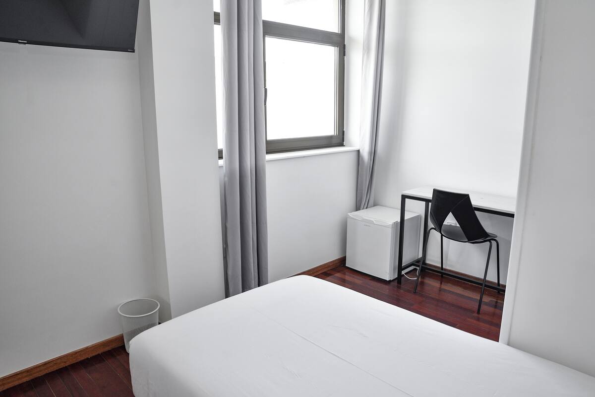 Quarto pequeno e limpo, com uma cama, escrivaninha, cadeira e janela com cortinas.