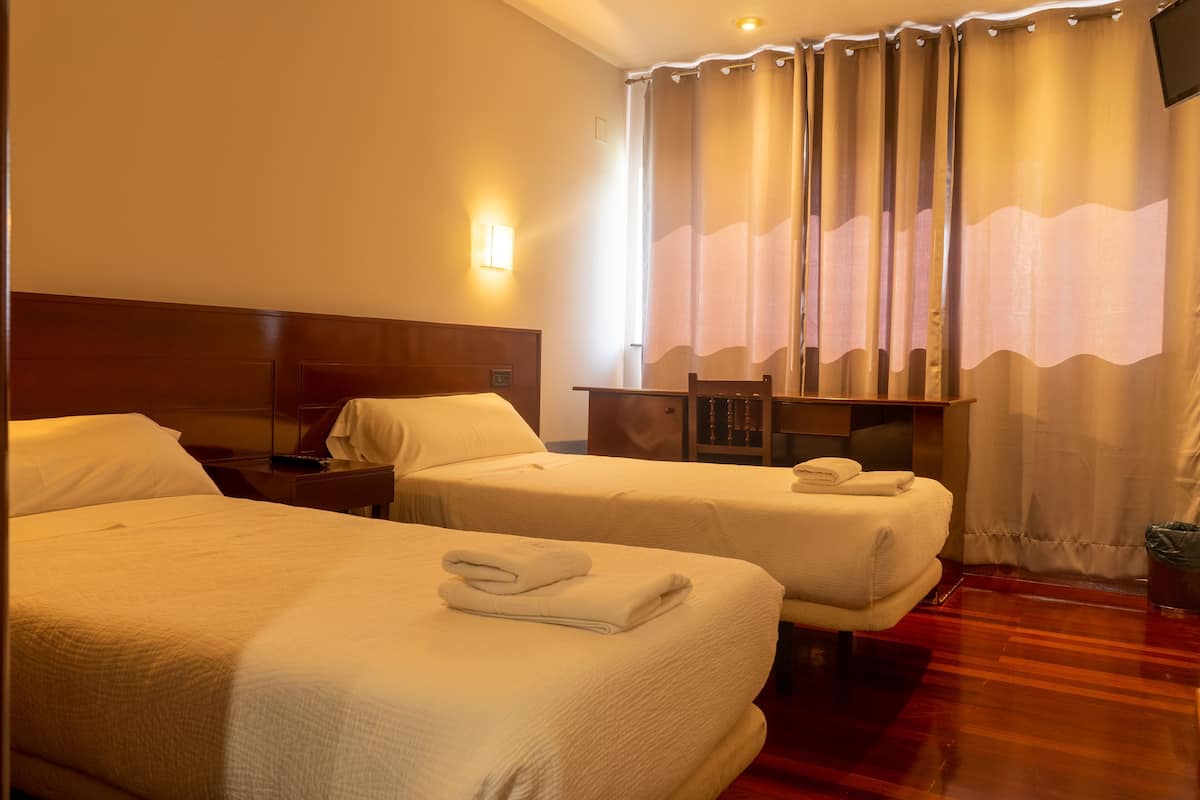 Quarto de hotel com duas camas, uma mesa de cabeceira, uma luminária e uma janela com cortinas.