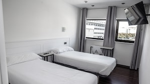 Habitación de hotel con dos camas individuales, un televisor montado en la pared, un escritorio pequeño y una ventana con cortinas.