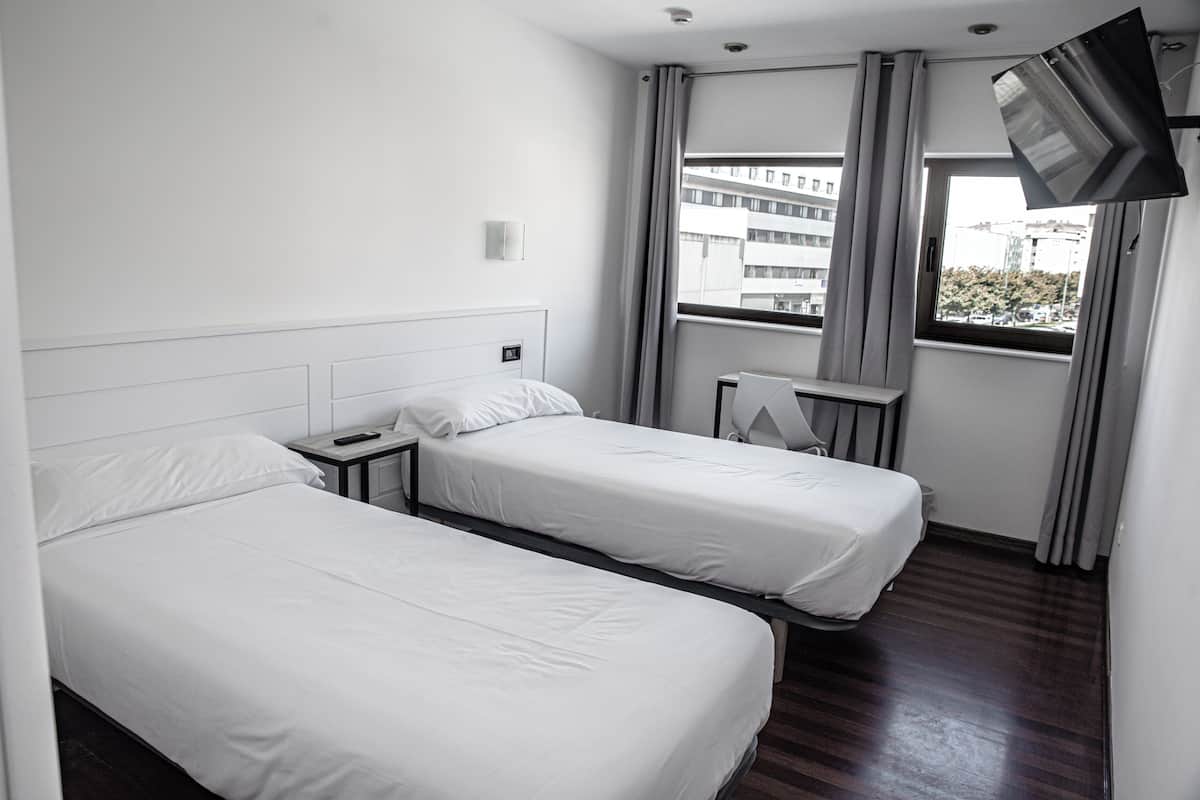 Quarto de hotel com duas camas de solteiro, televisão fixada na parede, uma secretária pequena e uma janela com cortinas.