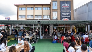 Un área de comedor al aire libre con mesas y sillas, un edificio con un letrero que dice 'CHIPS' al fondo y un corredor techado.