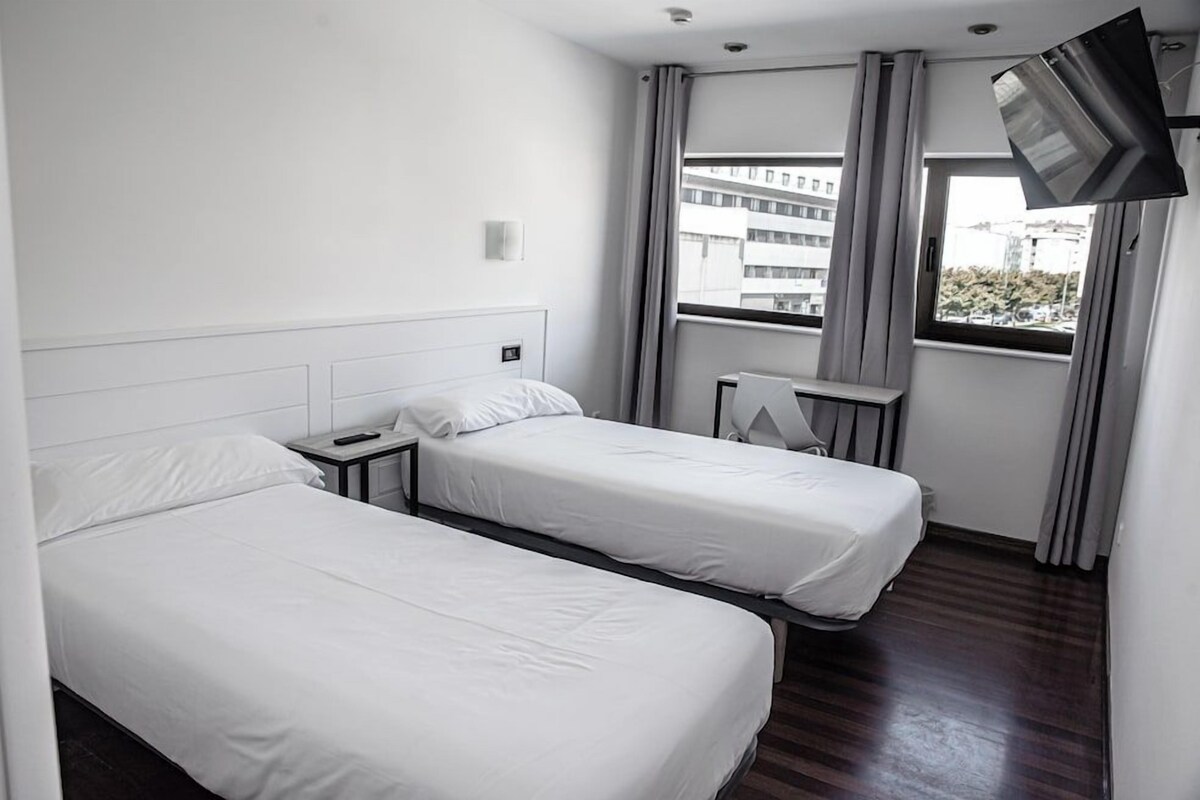 A hotel room with two single beds, a desk, a chair, a flat-screen TV, and a window with curtains.