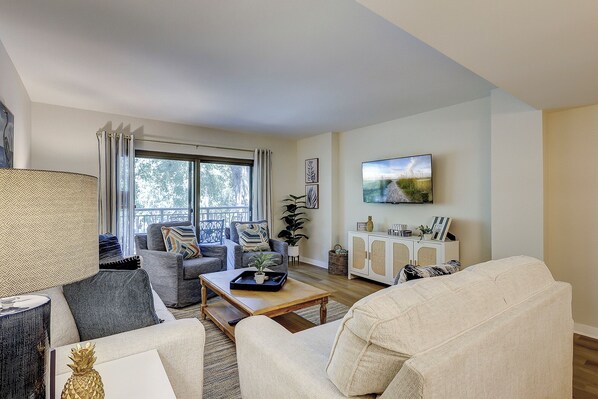 A living room with a sofa, armchairs, a coffee table, and a TV. There is a window with curtains, a plant, and a cabinet with decorative items. - Sunset Rentals | Ocean One 320 (Hilton Head Island)