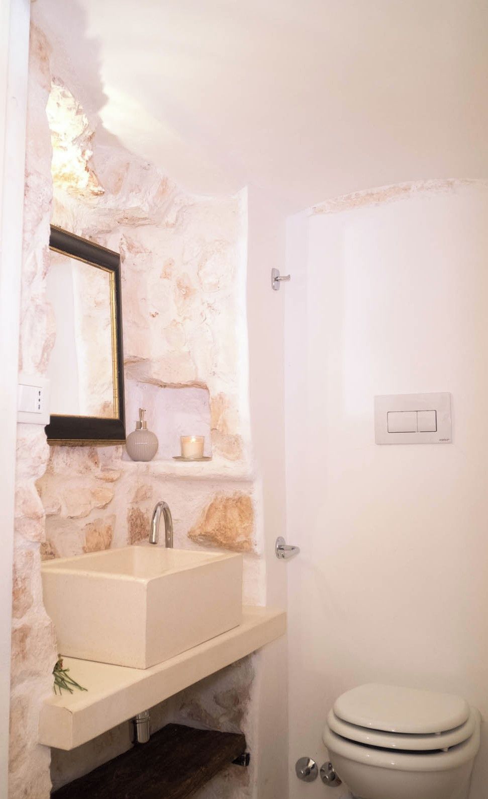 Un baño con pared de piedra, un lavamanos, un espejo, un inodoro y un interruptor de luz.