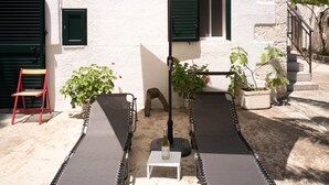 Um pátio com um edifício branco, um guarda-sol bege, duas cadeiras de descanso e plantas em vasos.