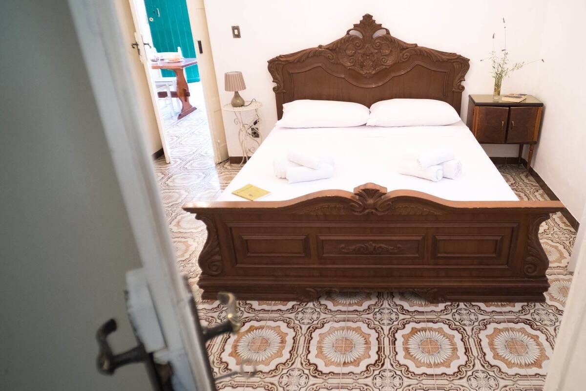 Uma cama bem arrumada com lençóis brancos, cabeceira de madeira e um chão estampado.