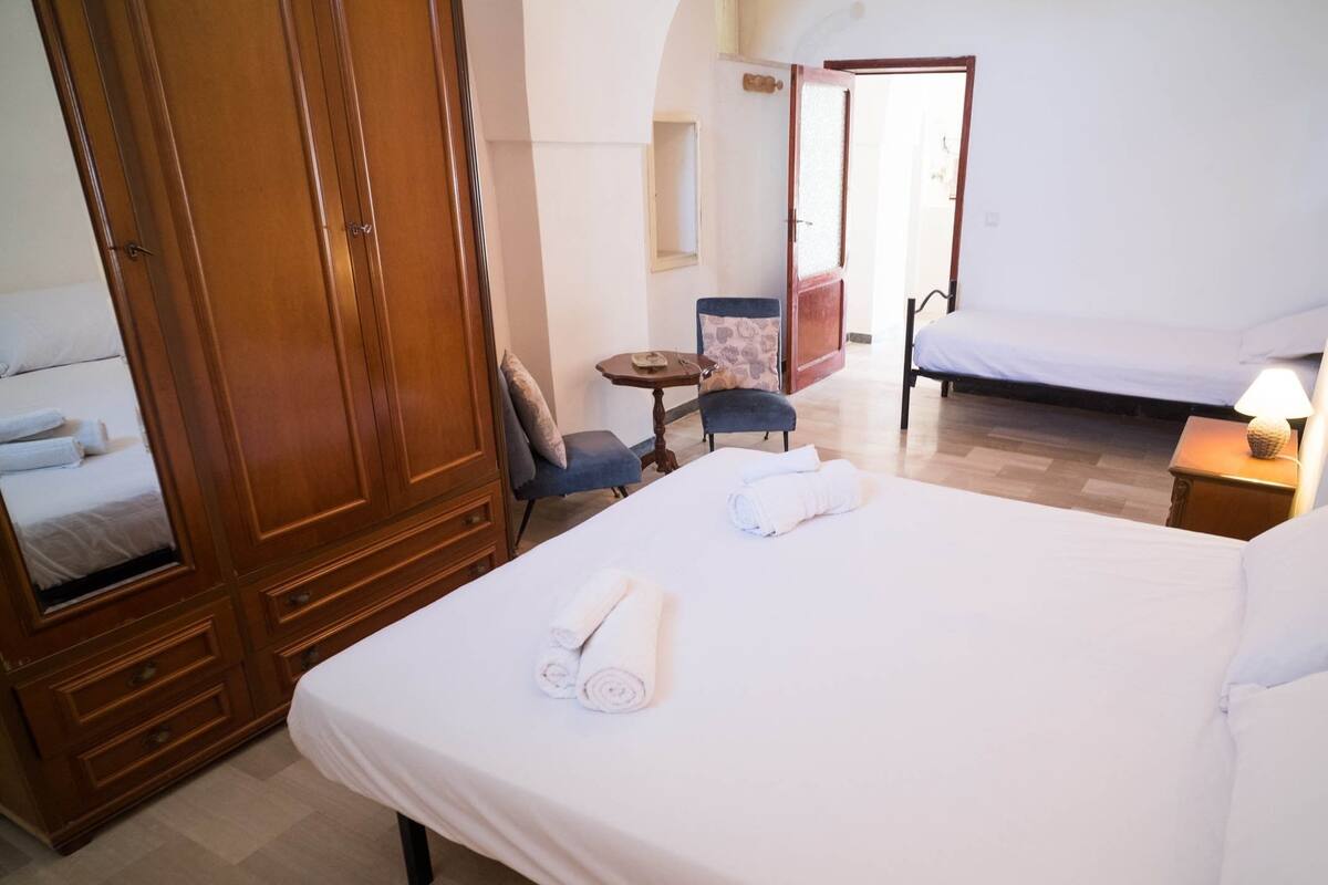 Quarto de hotel com uma cama grande, um guarda-roupa de madeira, uma mesinha, uma cadeira e uma escrivaninha com uma luminária.
