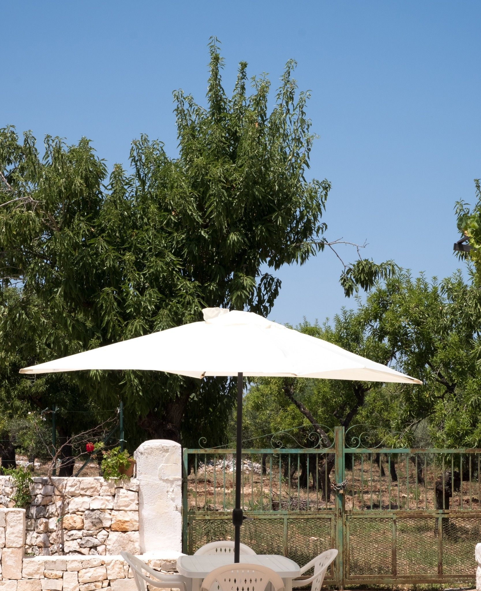An outdoor seating area with a table and chairs under a large umbrella, surrounded by stone walls and a metal fence.
