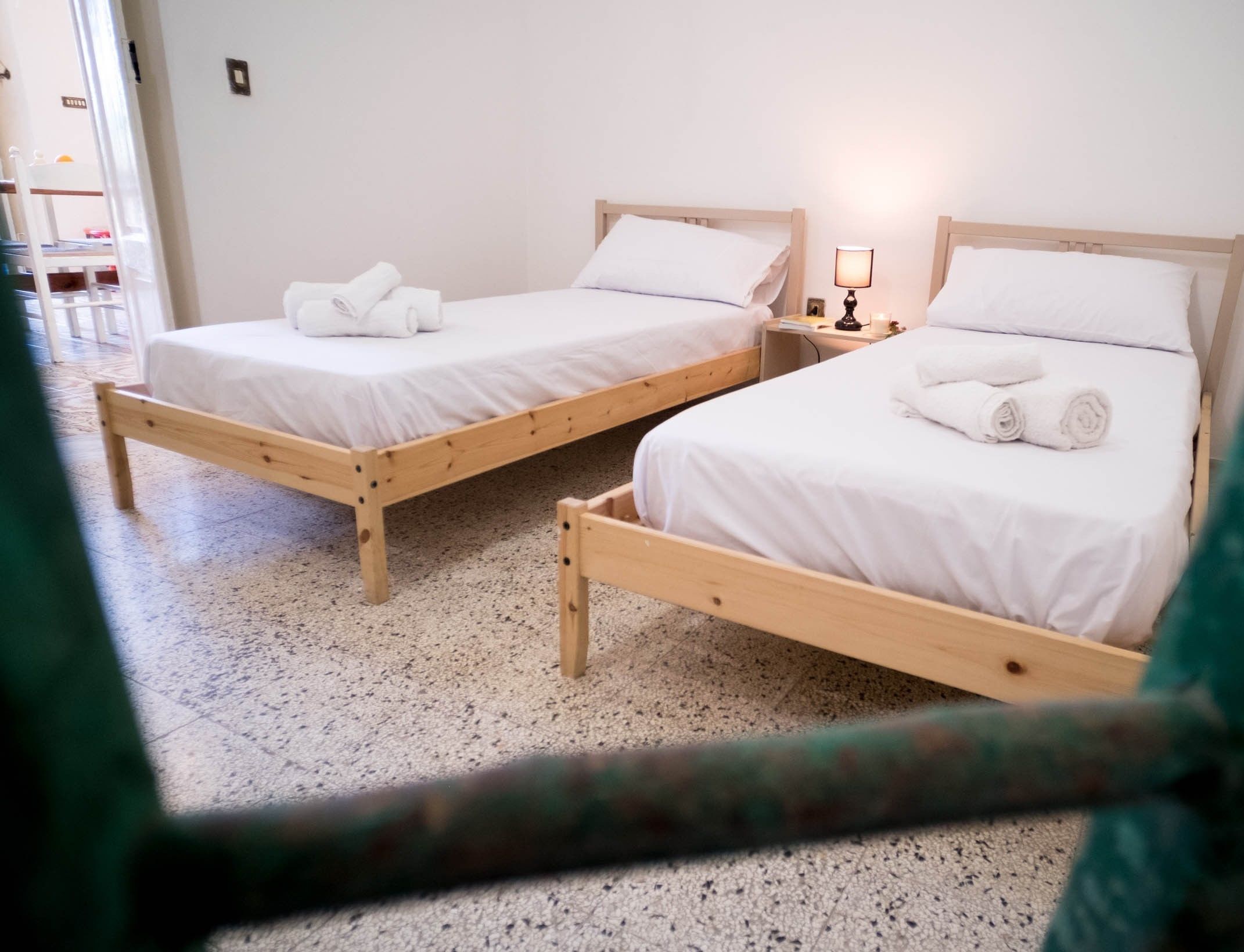 Dos camas de madera con sábanas y toallas blancas, una mesita de noche con una lámpara y un piso moteado.