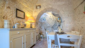 A rustic kitchen with stone walls, a wooden table, white chairs, and a cabinet with kitchenware. - La Lamia 1864 Ostuni by Typney (Ostuni)