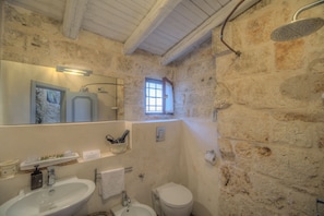 A bathroom with a stone wall, a shower, a sink, a toilet, and a window.