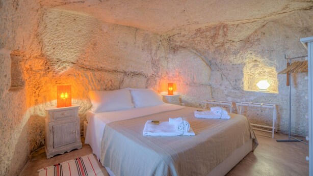 A bedroom with a stone wall, a bed, a nightstand, a lamp, and a wooden hanger rack. - La Lamia 1864 Ostuni by Typney (Ostuni)