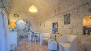 A rustic stone interior with a round table and white chairs, a small shelf with books and a radio, and a stone fireplace. - La Lamia 1864 Ostuni by Typney (Ostuni)