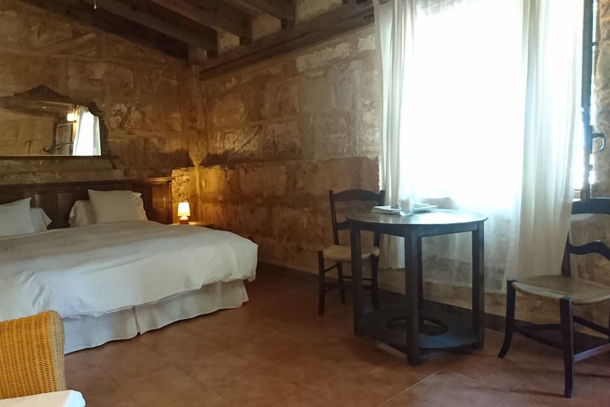 Una habitación rústica con una cama grande, una mesita redonda con sillas, una lámpara y un ventanal con cortinas.