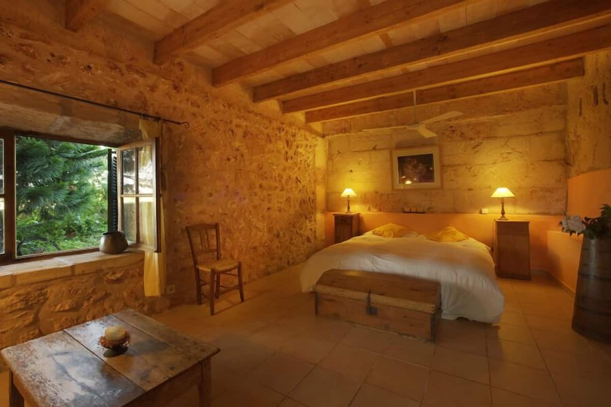 Una habitación rústica con cama de madera, paredes de piedra, ventana con vistas a follaje y dos lámparas de noche.