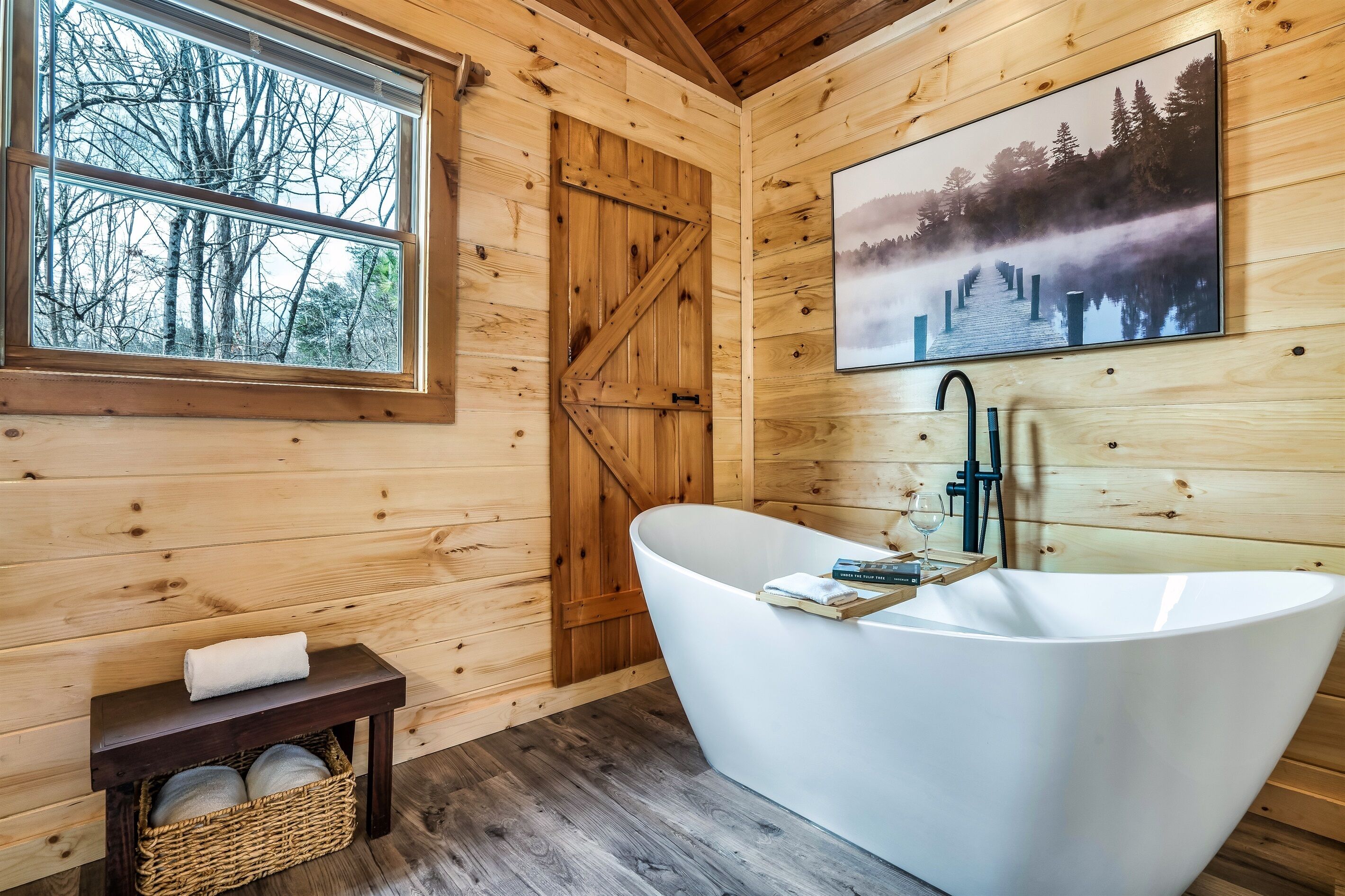 A wooden cabin bathroom with a freestanding bathtub, a window with a view of trees, and a painting of a forest scene.