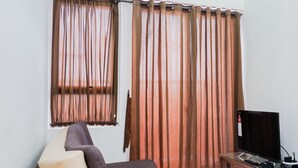 A room with a single reclining chair, a television on a wooden stand, and curtains on the windows. - Best Price 2BR at Taman Melati Margonda Apartment (Depok)