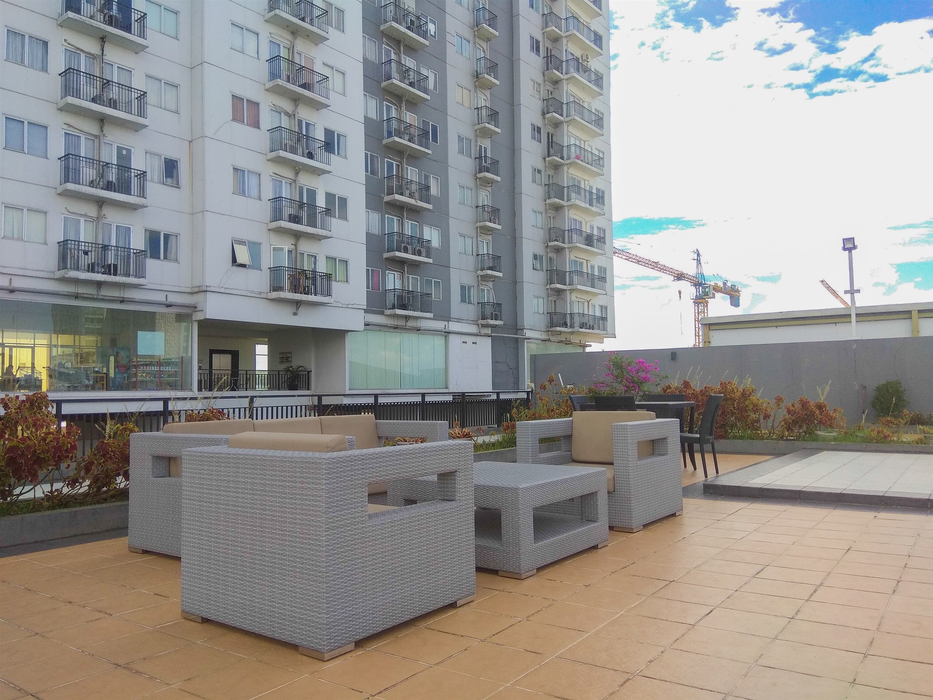 A modern outdoor seating area with wicker furniture, a building with multiple balconies in the background, and a construction crane visible.