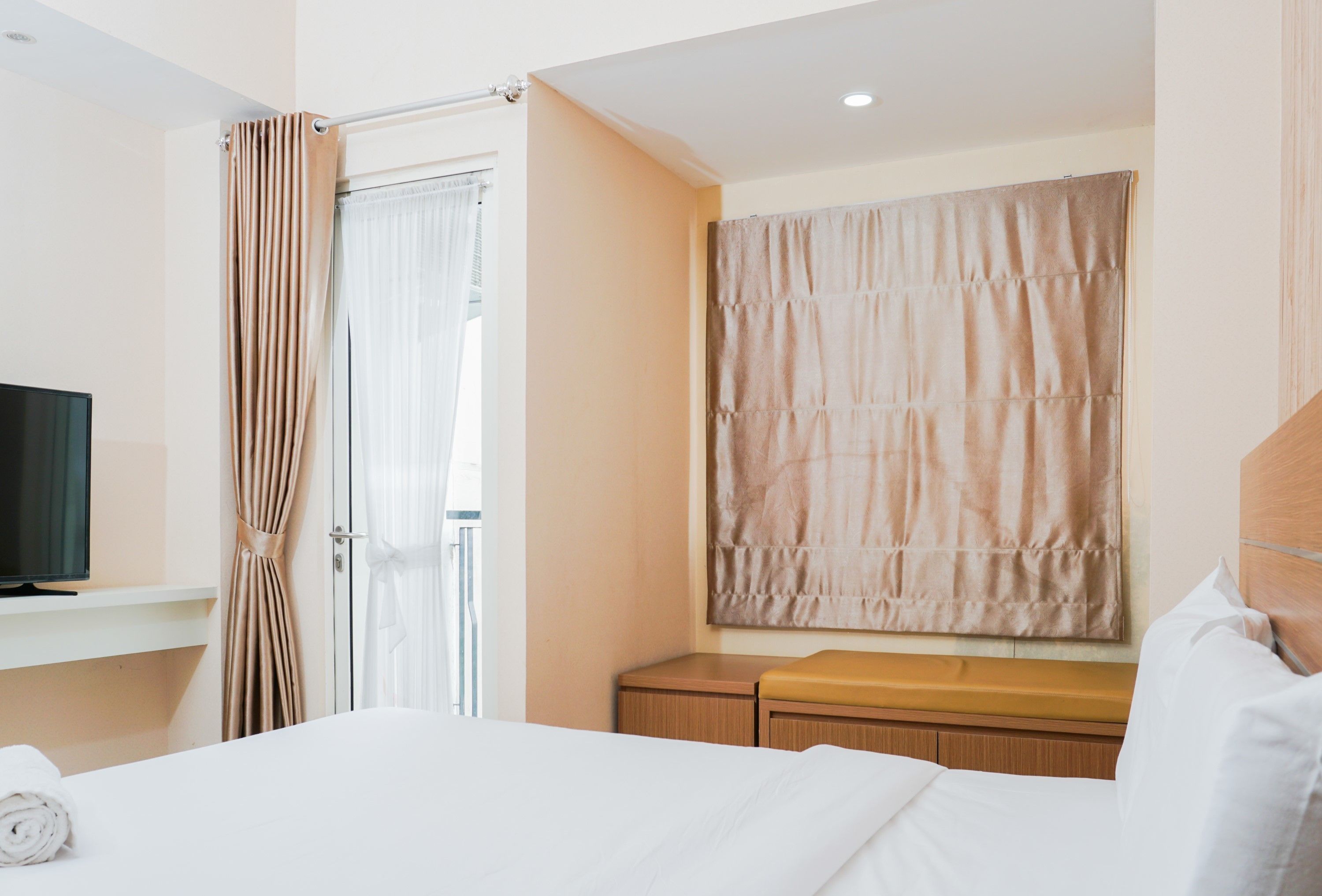 Habitación de hotel con cama, televisión, ventana con cortinas, un banco de madera y una pequeña mesa de madera.
