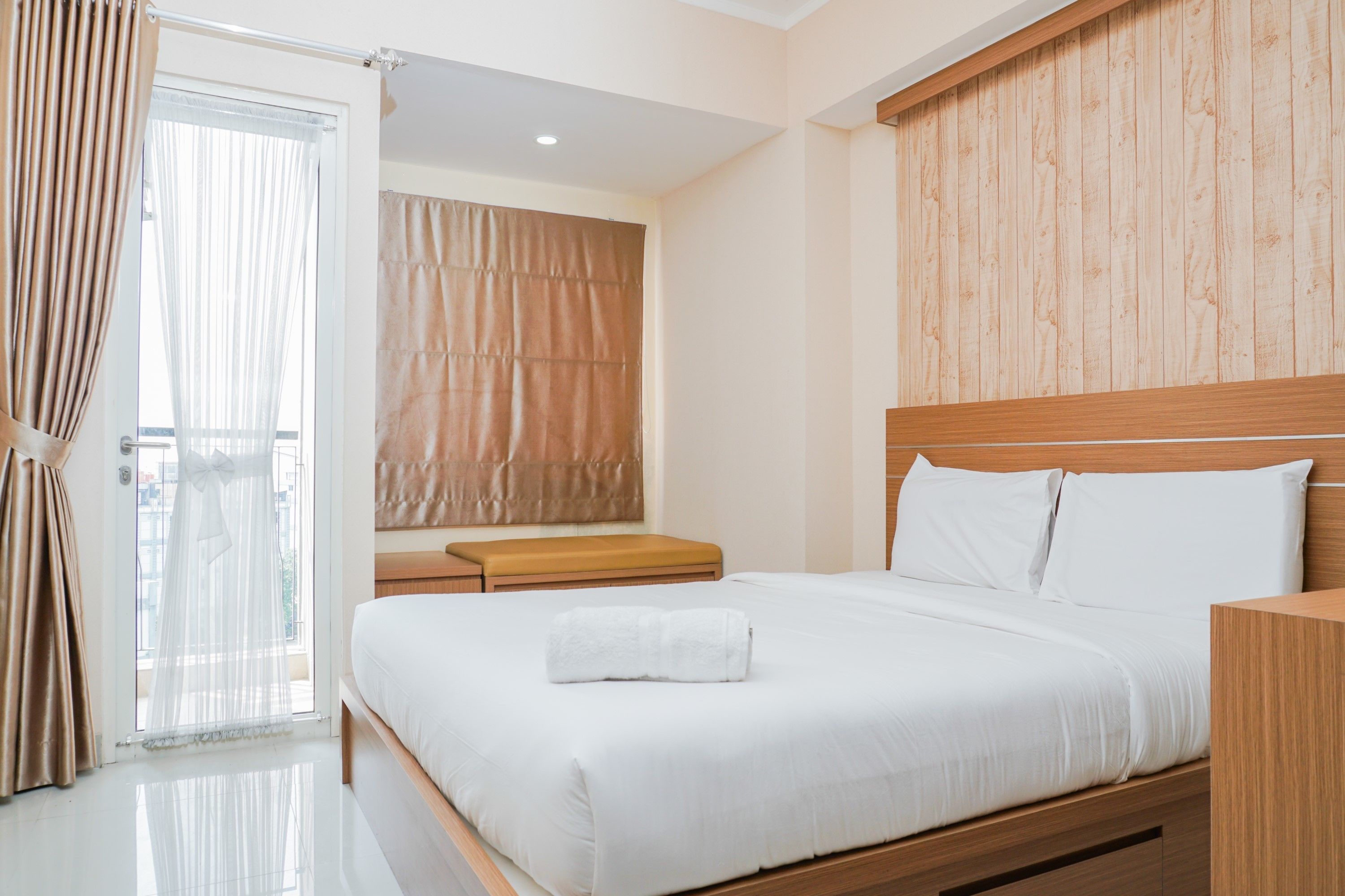 A modern hotel room with a wooden headboard, a large bed with white linens, a bench at the foot of the bed, and a view of the city through the window.
