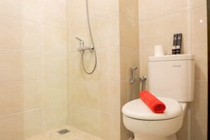 A bathroom with beige tiles, a shower, a toilet with a red towel, and a toilet paper holder. - Elegant Studio Mustika Golf Residence Apartment (Cikarang)
