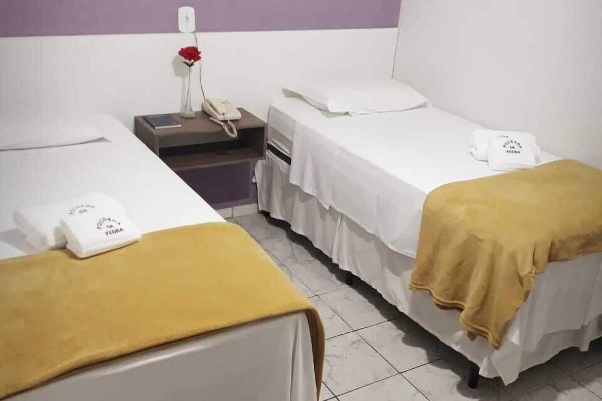 Une chambre d’hôtel avec deux lits simples, chacun recouvert d’un drap blanc et d’une couverture jaune. On y trouve une petite table avec un téléphone et un vase contenant une fleur rouge. Les murs sont peints en blanc et en violet, et le sol est carrelé.
