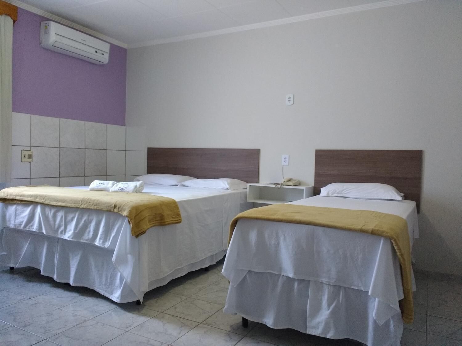 A hotel room with two beds, each with a white bedspread and a yellow blanket. There is a wall-mounted air conditioner and a phone on a nightstand.