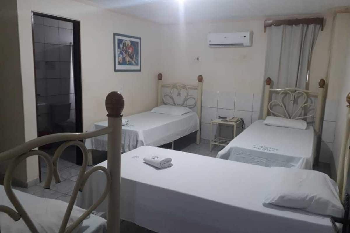 Une chambre d’hôtel avec trois lits simples, une petite table de chevet avec une lampe, un climatiseur fixé au mur et une porte donnant sur une salle de bain.