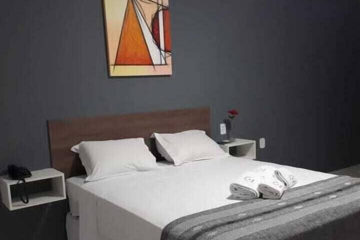 Un lit bien fait, avec du linge de lit blanc et un plaid gris, une table de chevet avec un téléphone et un vase contenant une fleur, et un tableau représentant une tente accroché au mur.