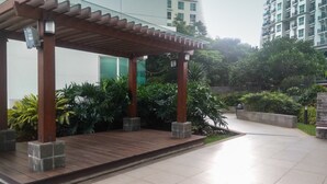 Un espace extérieur abrité doté d’une terrasse en bois, entouré de verdure et avec un bâtiment moderne en arrière-plan.
