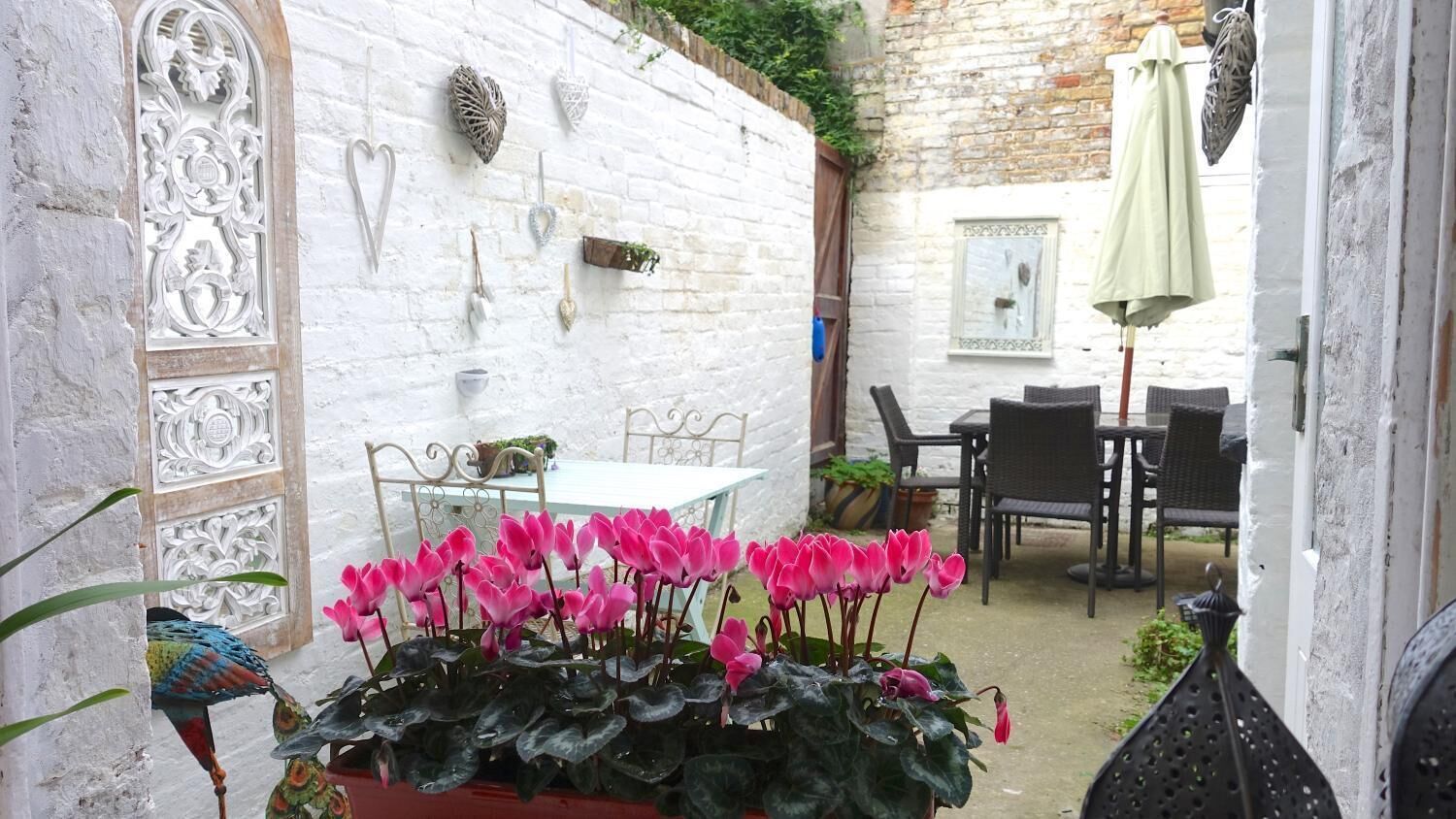 Un mur en briques blanches agrémenté d’éléments décoratifs en métal, une table avec des chaises et un pot de fleurs contenant des fleurs roses.